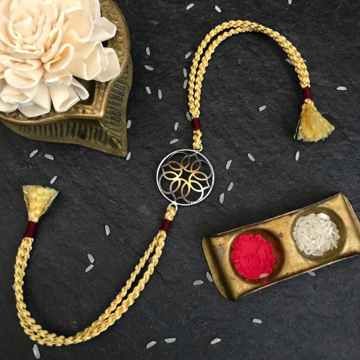 Handcrafted Rakhi with Traditional Braided Thread featuring a gold and red design, tassels, and a decorative gold dish with rice.