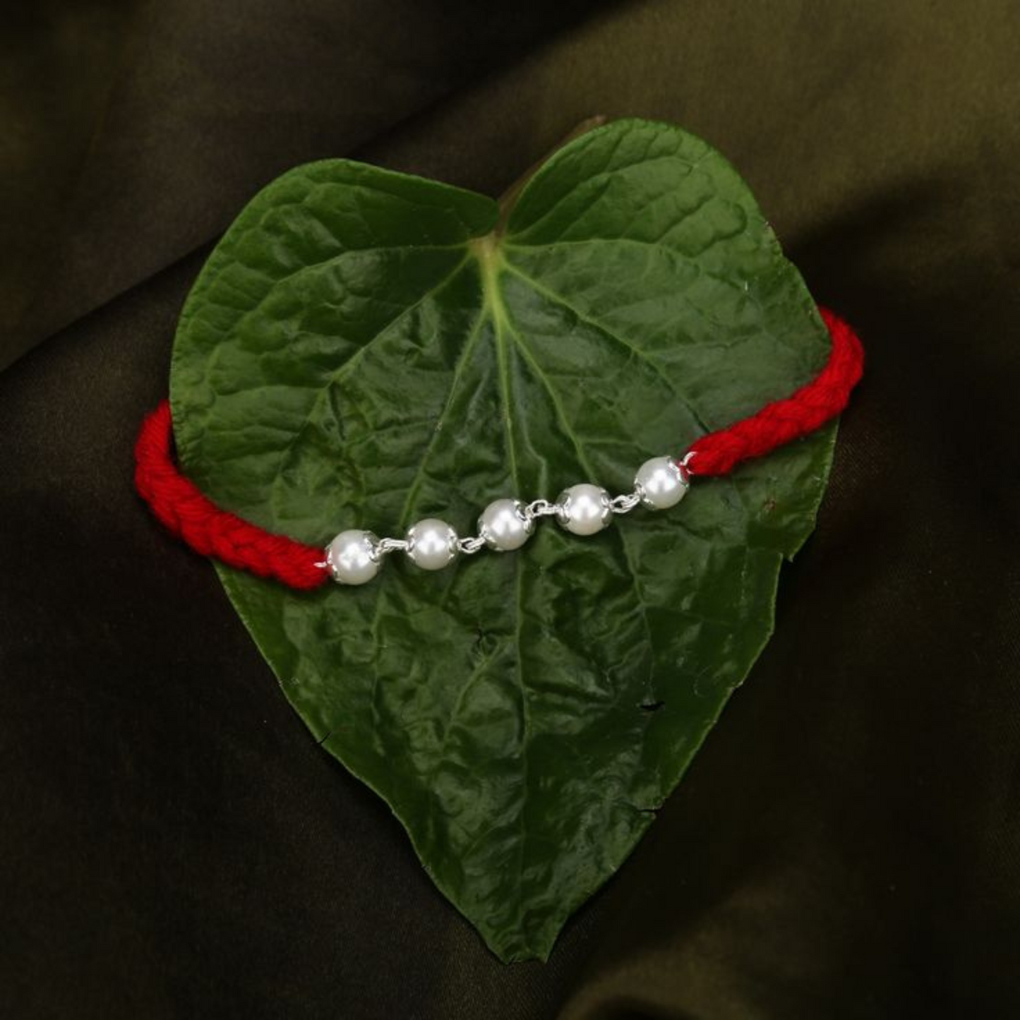 Elegant Pearl Rakhi with Red Thread by Bliss Jouels, displayed on a leaf. Features a red string and lustrous pearls, symbolizing purity and strength.