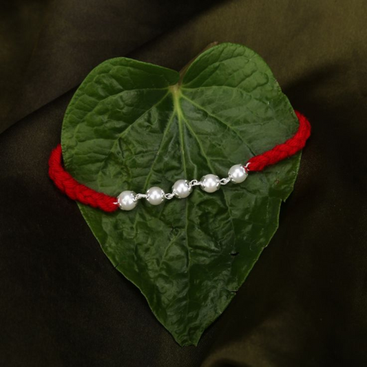 Elegant Pearl Rakhi with Red Thread by Bliss Jouels, displayed on a leaf. Features a red string and lustrous pearls, symbolizing purity and strength.