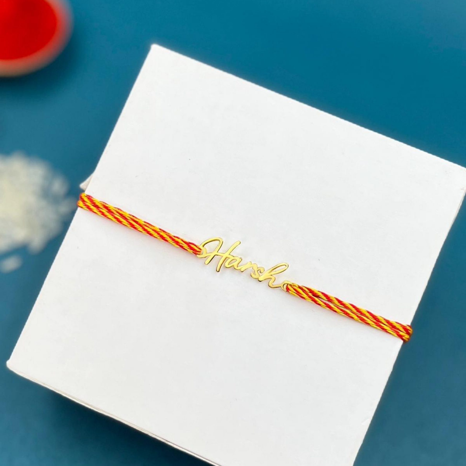 Alt text: Bliss Jouels Customized Name Rakhi featuring a white box with a yellow and red string, showcasing a gold name.